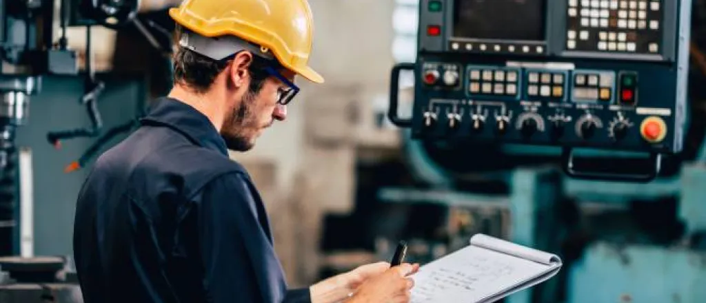 A team member wearing a blue jumpsuit and a yellow hard hat stands next to an automation machine, writing on a notepad.