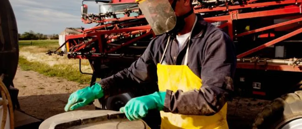 A person wearing a protective shield, a yellow protective apron, green protective gloves, and a mask is opening a container.
