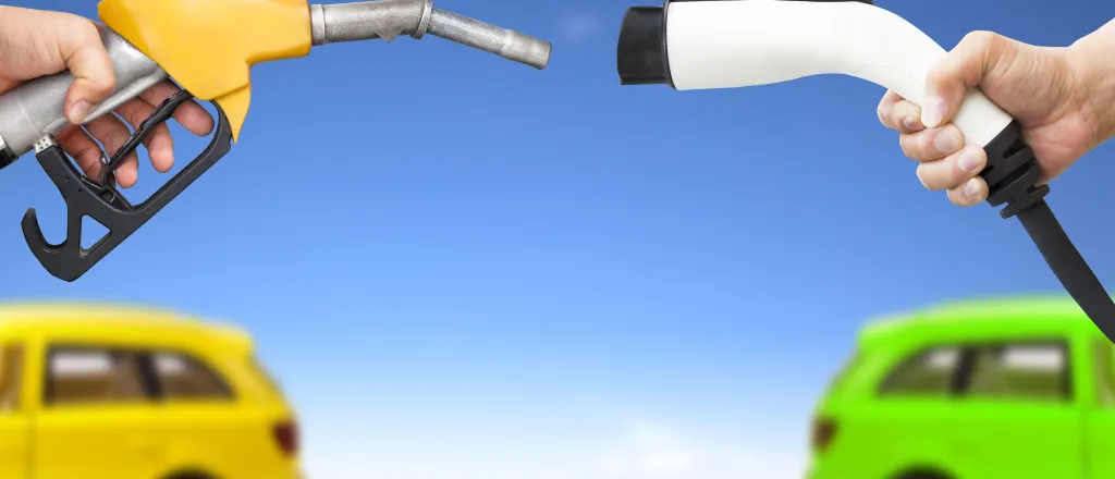 Hands holding a gasoline fuel nozzle and electric charging cable in the foregrounds, with two vehicles in the background.