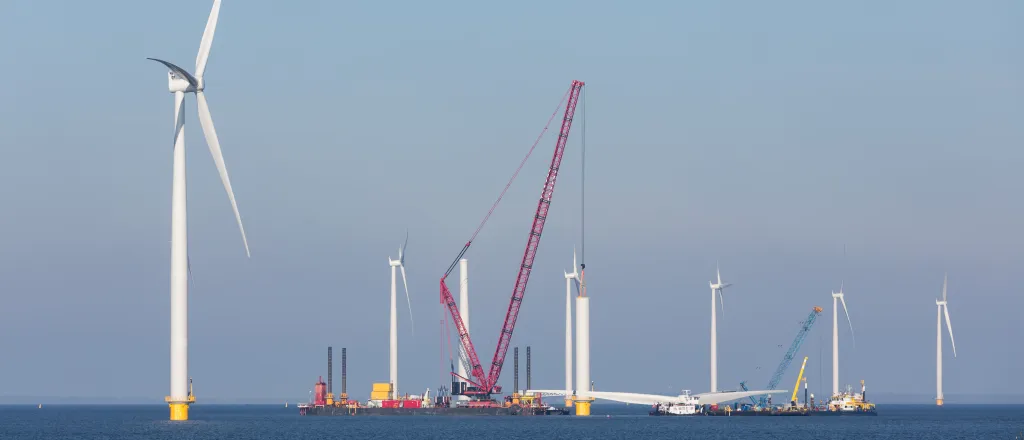 Wind turbines under construction in the ocean. A crane ship is in place to lift a blade assembly.