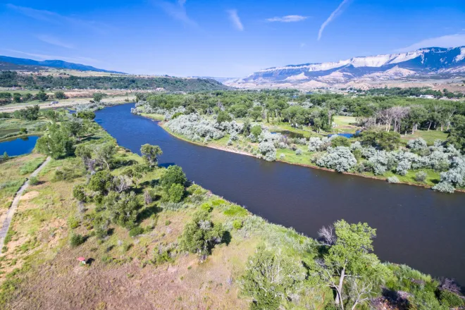 Aerial view of the Colorado River surrounded by trees and low vegetation with mountains and blue sky in the background.