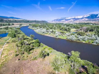 Aerial view of the Colorado River surrounded by trees and low vegetation with mountains and blue sky in the background.