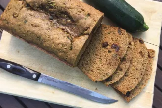 Overhead view of a partially sliced loaf of zucchini bread resting on a cutting board. A knife and whole zucchini rest on either side of the bread.